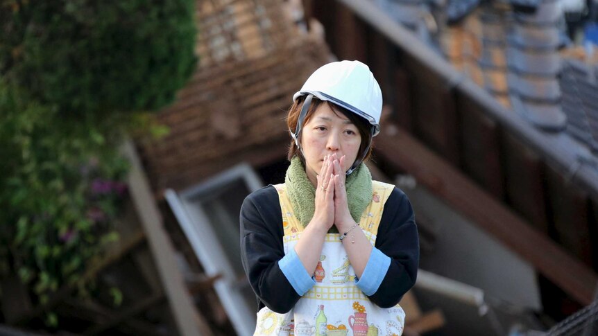 A woman reacts in front of a collapsed house