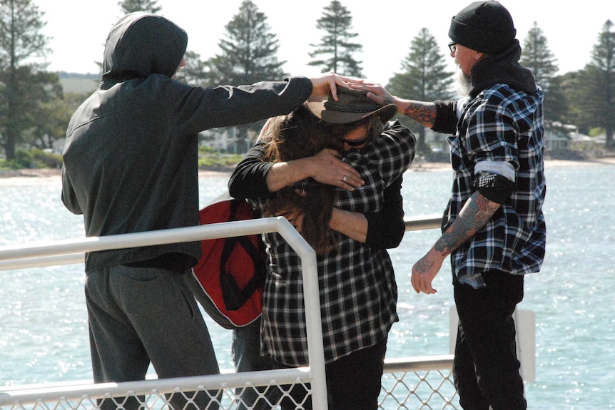 A man hugs a small woman while two men hold his brown hat on his head to stop it blowing away in the wind