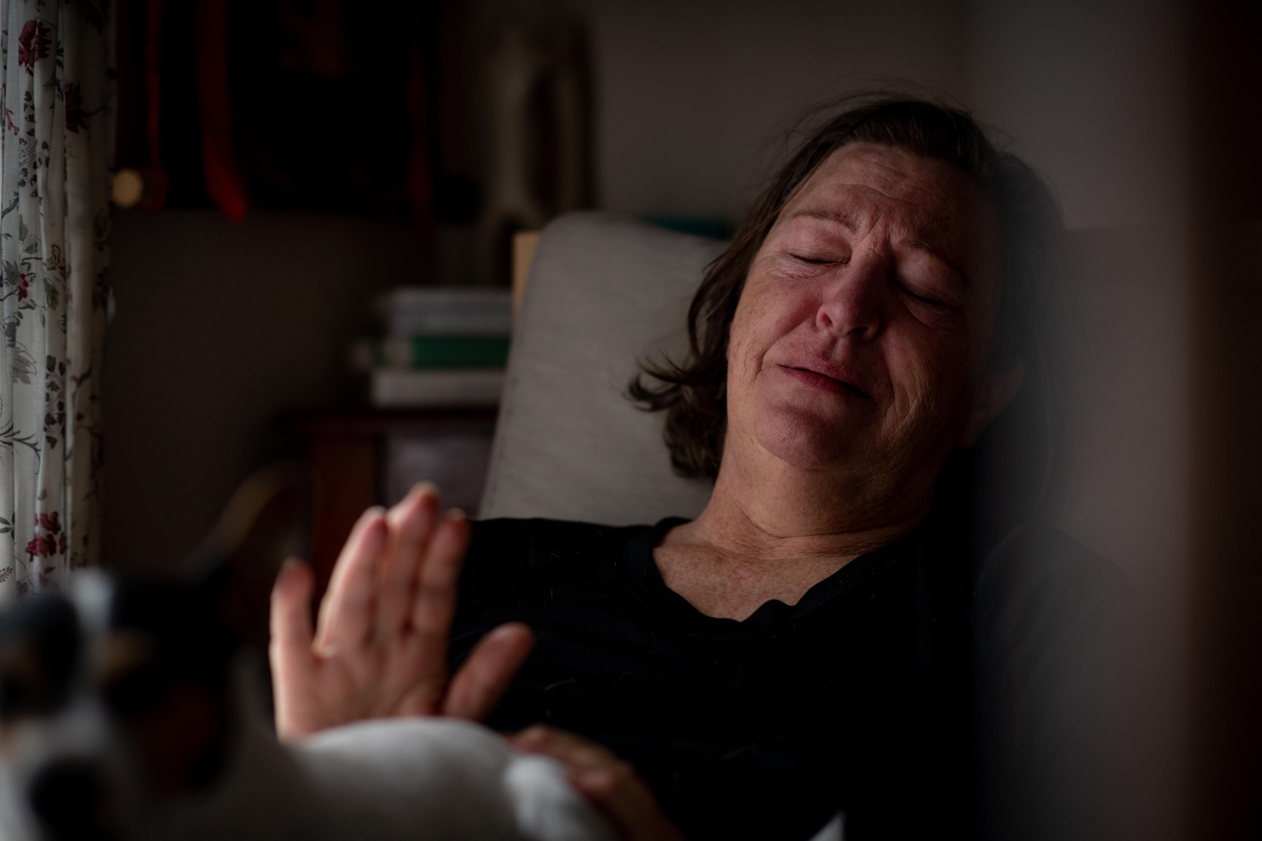 A middle-aged white woman lying in bed looking sad and exhausted. She is lifting her hand up