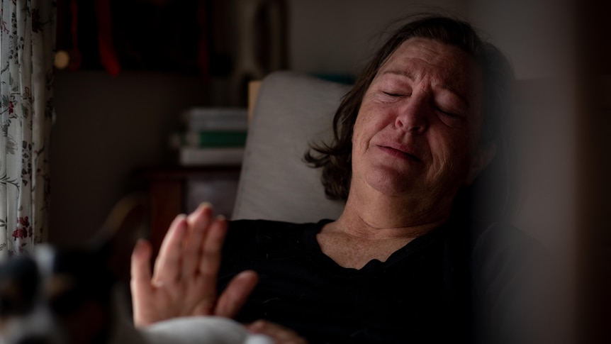 A middle-aged white woman lying in bed looking sad and exhausted. She is lifting her hand up