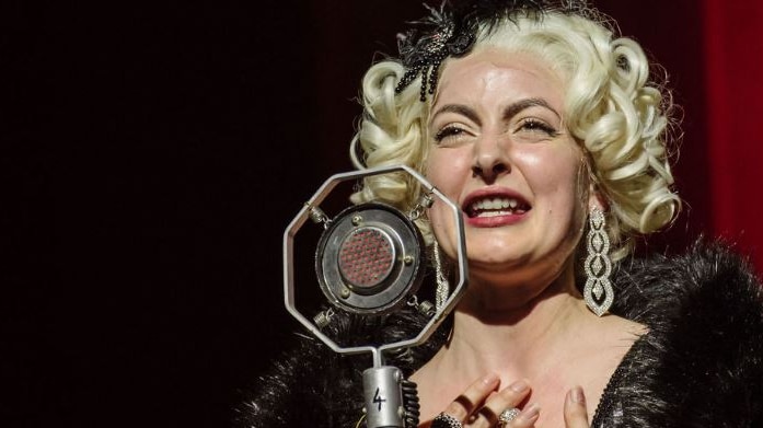 An actress in a blond wig talks into a microphone as Lina Lamont on stage.