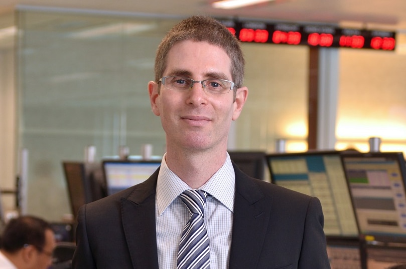 A man in suit standing in front of computers at a financial trading desk.