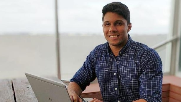 A young man, Mohamed Nizamdeen, sits at a table with a laptop open in front of him. He is smiling at the camera.