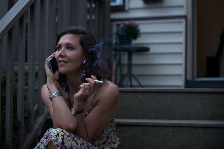 Colour close-up still of Maggie Gyllenhaal talking on phone and smoking on porch steps in 2018 film The Kindergarten Teacher.