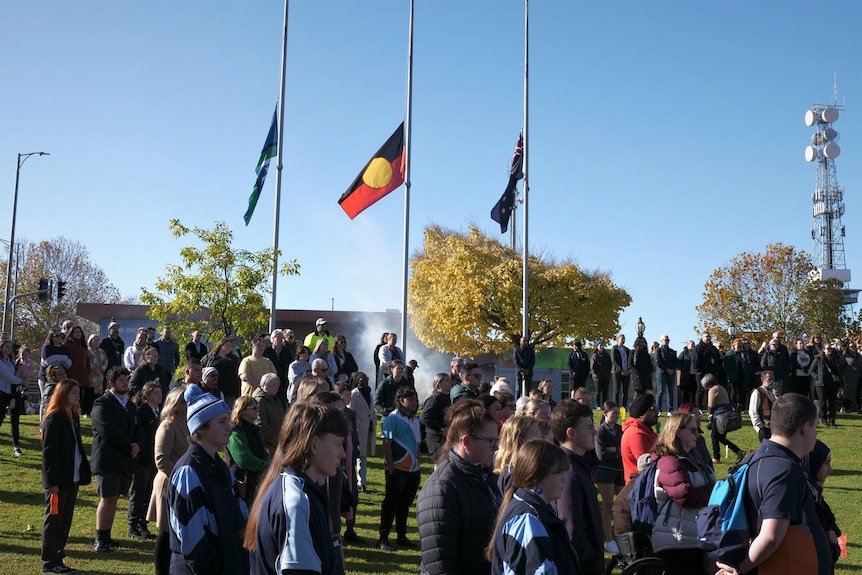 A group of people gathering outside, there are three flag poles with flags flying half mast. 