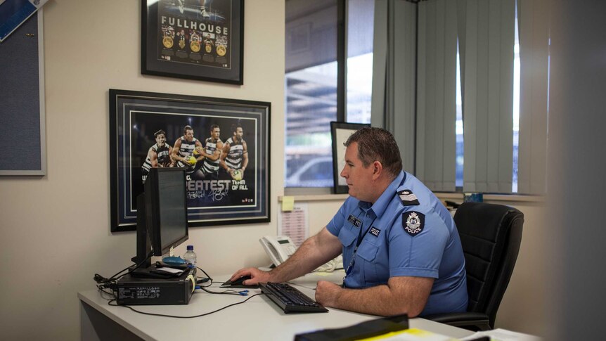 A police officer, wearing a blue shirt, sits at a desk in his office