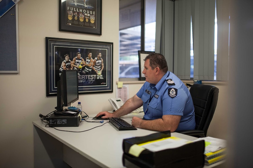 A police officer, wearing a blue shirt, sits at a desk in his office