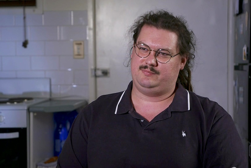 A man with long dark hair wears glasses and a black collared shirt.