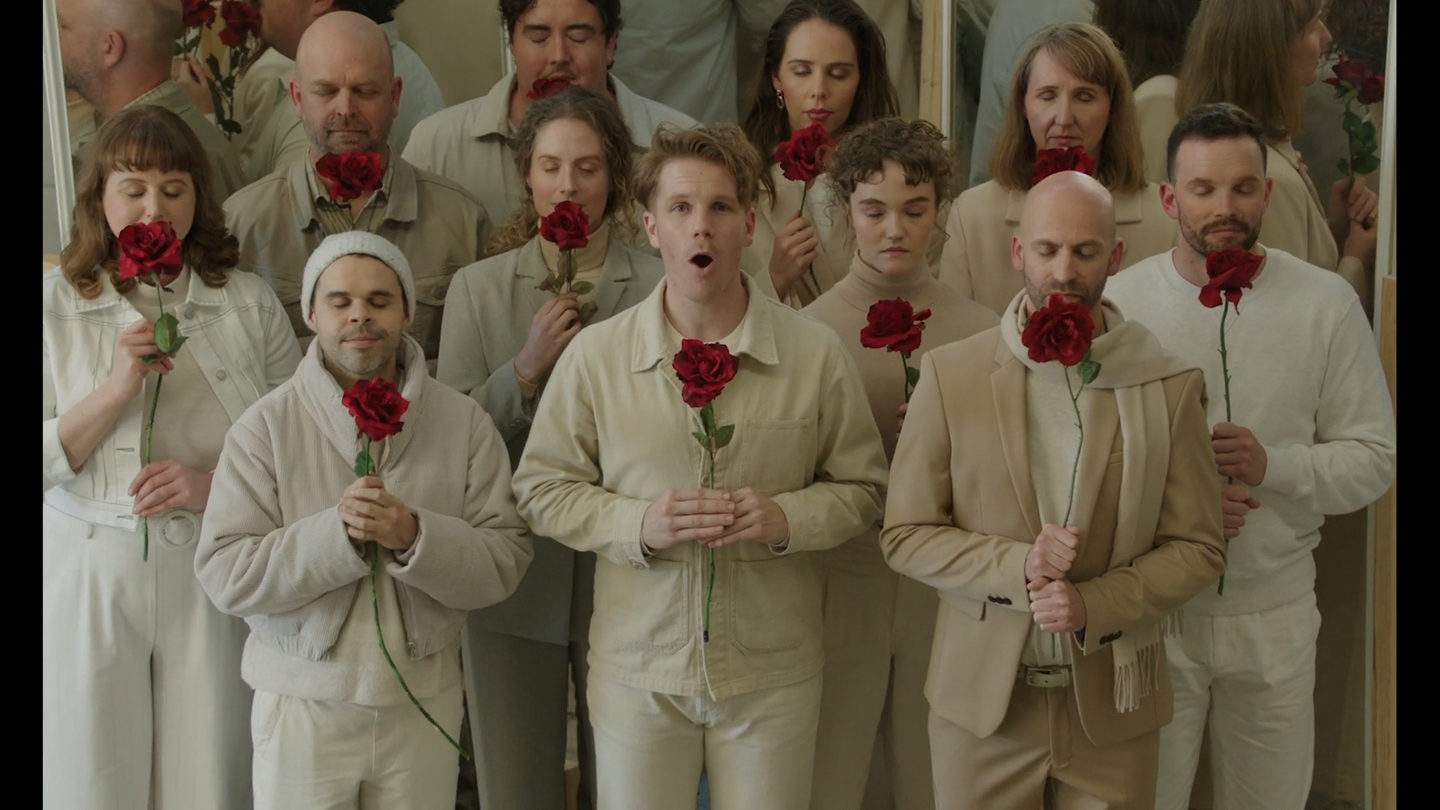 A group of singers dressed in white and beige look at the camera with large red flowers in their hands. Most of them have their eyes closed while singing.