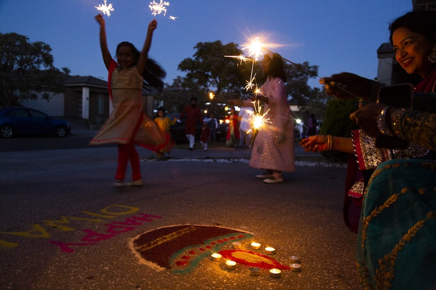 Two girls wearing traditional Indian clothes twirl while holding sparklers in front of a clay lamp and Happy Diwali drawing.