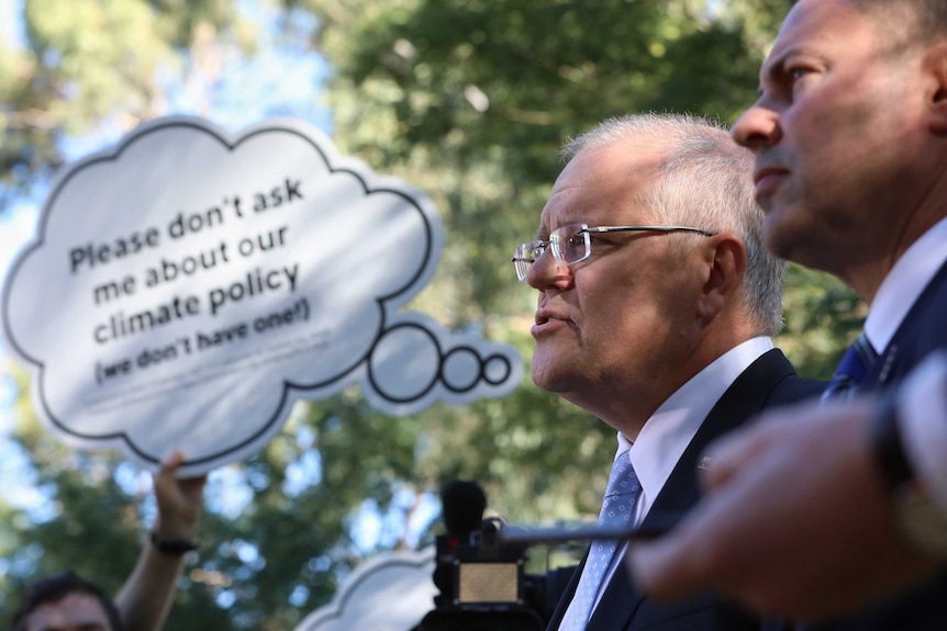 A protester holds a "please don't ask me about our climate policy" speech bubble alongside Scott Morrison and Josh Frydenberg