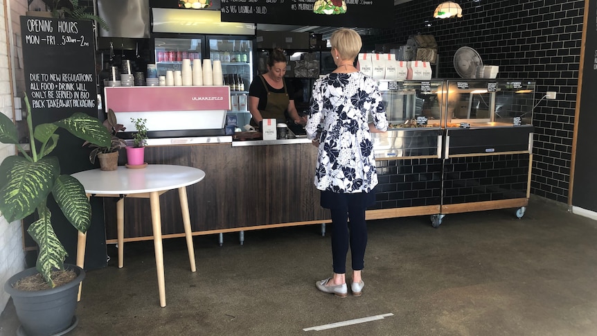 A woman stands in a cafe, with a woman serving her behind the counter.