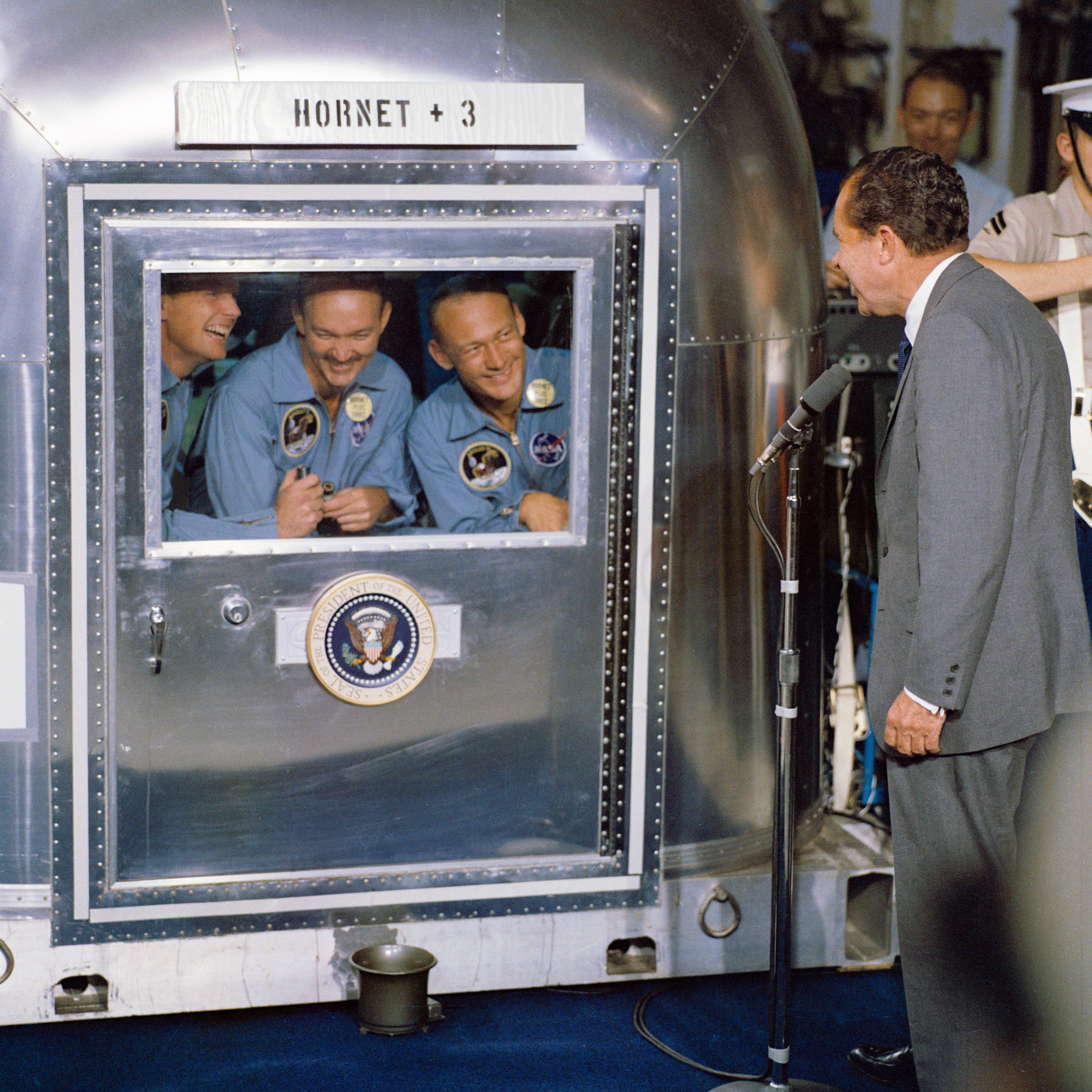 Foto de la década de 1970 que muestra una caja plateada con una ventana; Tres hombres sonrientes miran hacia afuera mientras un hombre con traje mira hacia adentro.