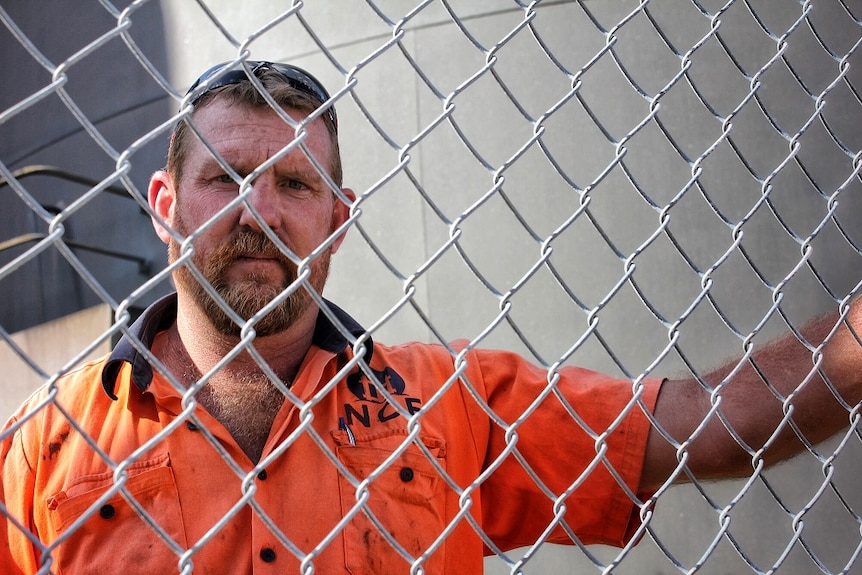 A man in an orange high-viz shirt standing behind a wire mesh fence, looking at the camera.