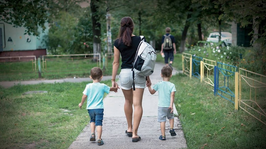 A woman wearing a backpack holds hands with two small children as they all walk down a footpath.