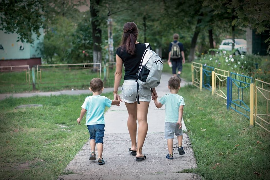 A woman wearing a backpack holds hands with two small children as they all walk down a footpath.