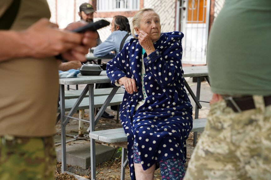 An elderly woman has a faraway look in her eye as she sits on a bench in a blue and white spotted dressing gown