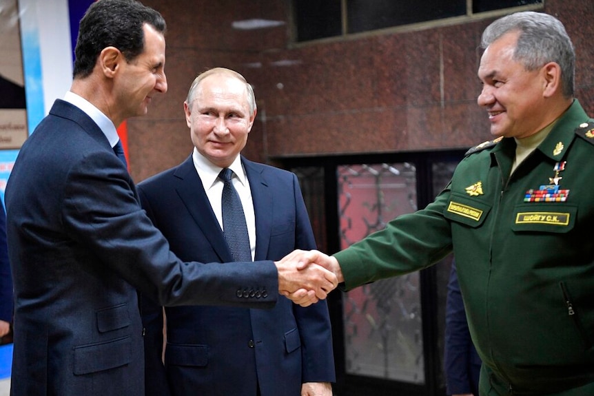 Vladimir Putin stands between Bashar Assad and Sergei Shoigu shaking hands in a dim room with stone walls.