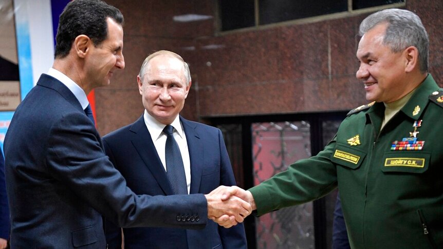 Vladimir Putin stands between Bashar Assad and Sergei Shoigu shaking hands in a dim room with stone walls.