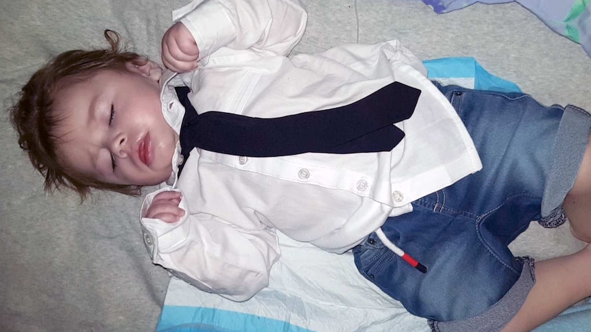 Nathaniel lying asleep on a quilt, and wearing a white shirt, black tie and blue denim shorts.