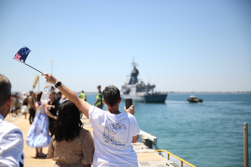 People gather on a jetty waving flags to farewell a navy ship