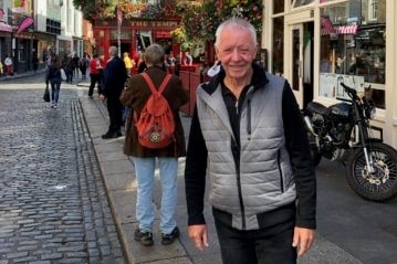 An elderly man stands in a cobble stone street and smiles to the camera.