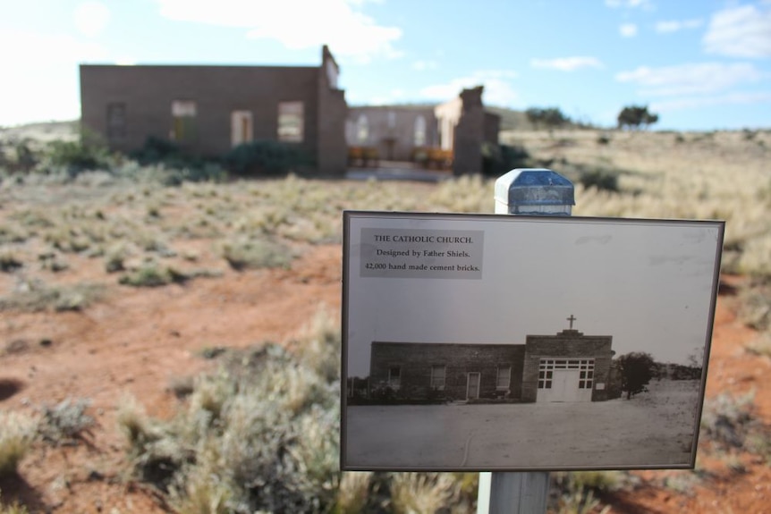 The ruins of an old brick building stand in the distance behind a sign depicting its former church glory