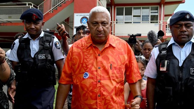 Fiji First leader Frank Bainimarama leaves a polling station wearing orange shirt and flanked by two police officers