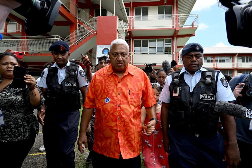 Fiji First leader Frank Bainimarama leaves a polling station wearing orange shirt and flanked by two police officers