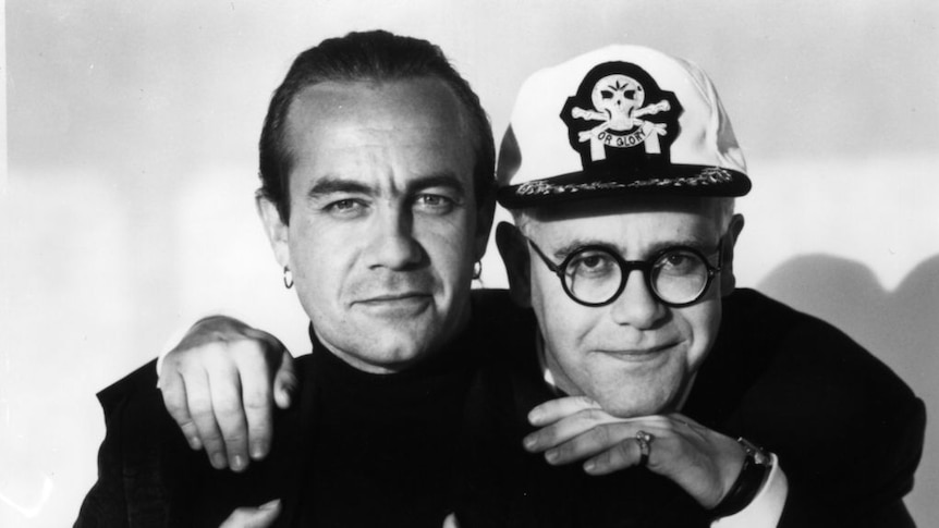 Two men smile at a camera, including one wearing a captain's hat.