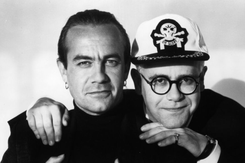 Two men smile at a camera, including one wearing a captain's hat.