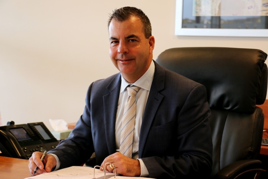 A man wearing a suit jacket and cream tie sits in a leather chair at a desk.