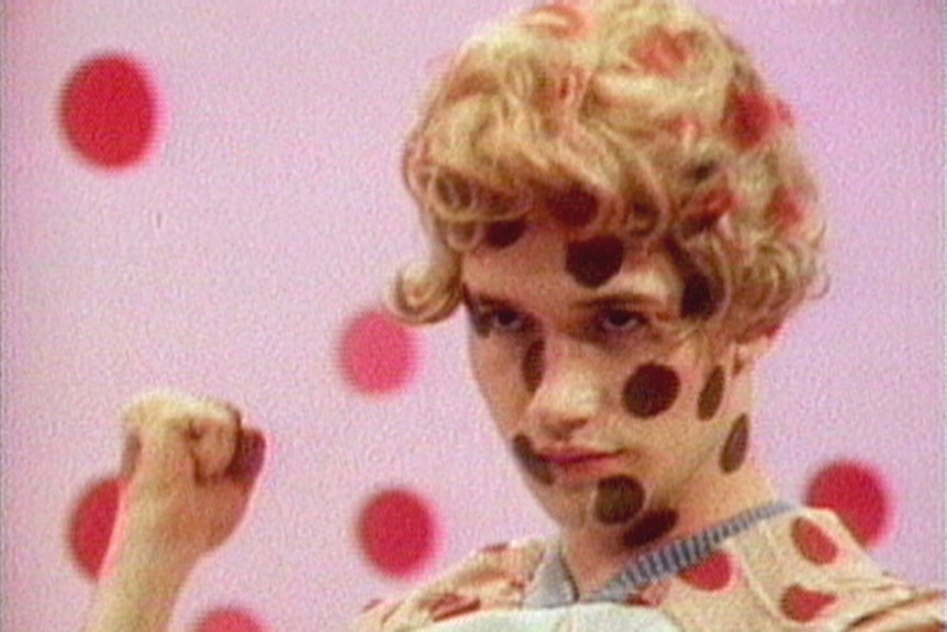 Colour still of a figure with blonde hair, clenching fist and covered in red polka dots, in front of pink wall also with dots.