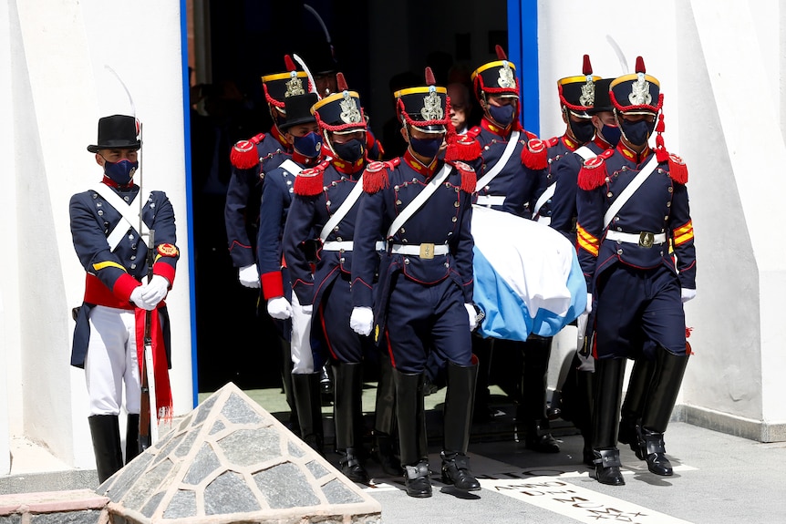 Guards in ceremonial uniform carry a coffin draped in the Argentinian flag through a doorway, as another officer stands guard. 