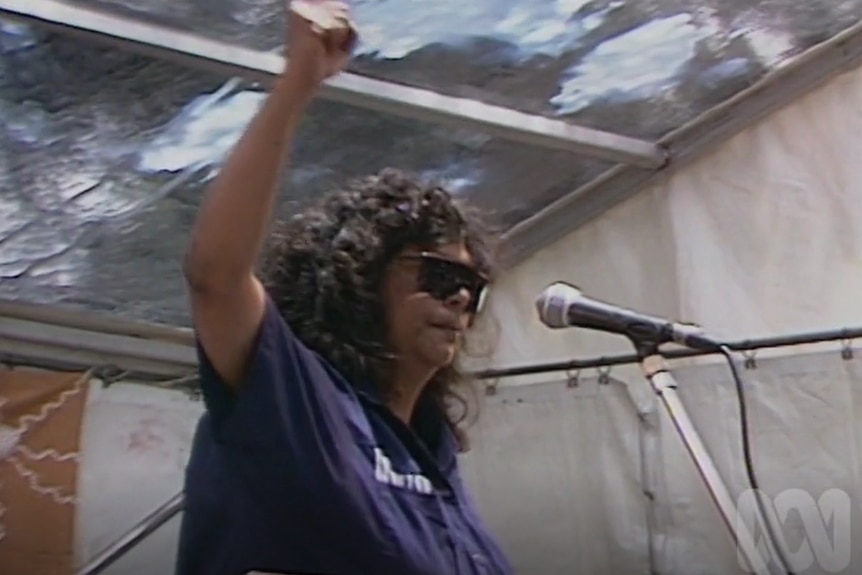 A woman in dark glasses stands by a microphone, her fist in the air.