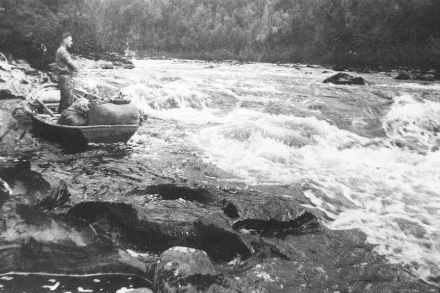 Historic image showing Huon piner standing in loaded wooden row-boat hauled onto rocks beside a river rapid