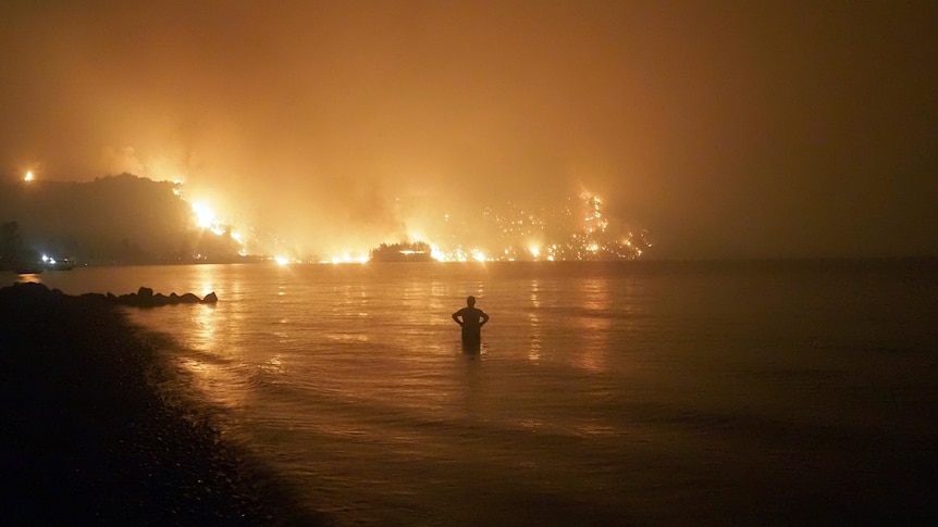 A man stands in water with flames in the distance