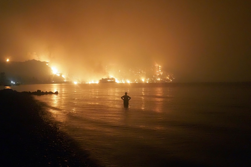 A man stands in water with flames in the distance