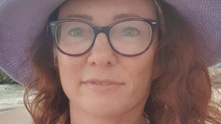 A close up of a woman with long wavy hair wearing a purple hat and thick rimmed glasses, looking into the camera.