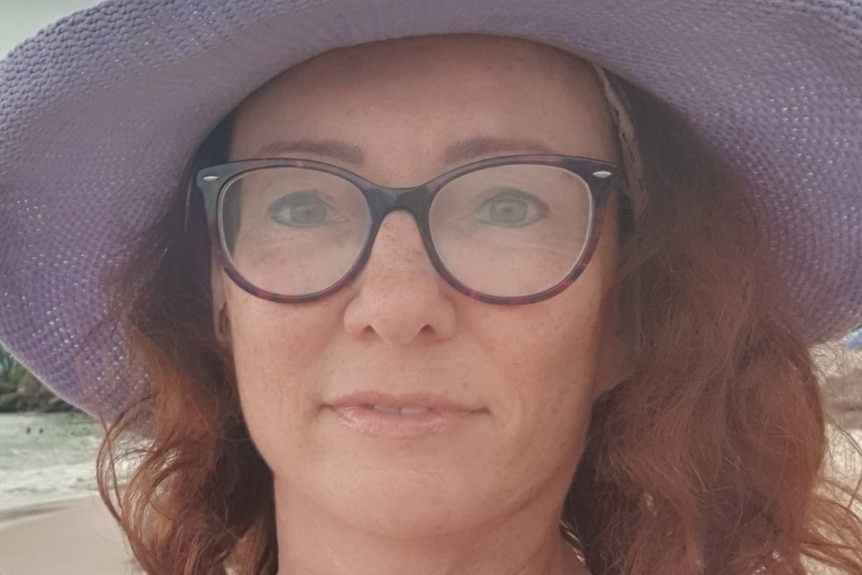 A close up of a woman with long wavy hair wearing a purple hat and thick rimmed glasses, looking into the camera.