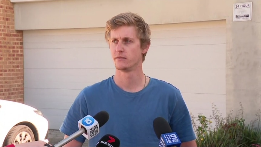 A young man with blonde hair wears a blue shirt and speaks to media