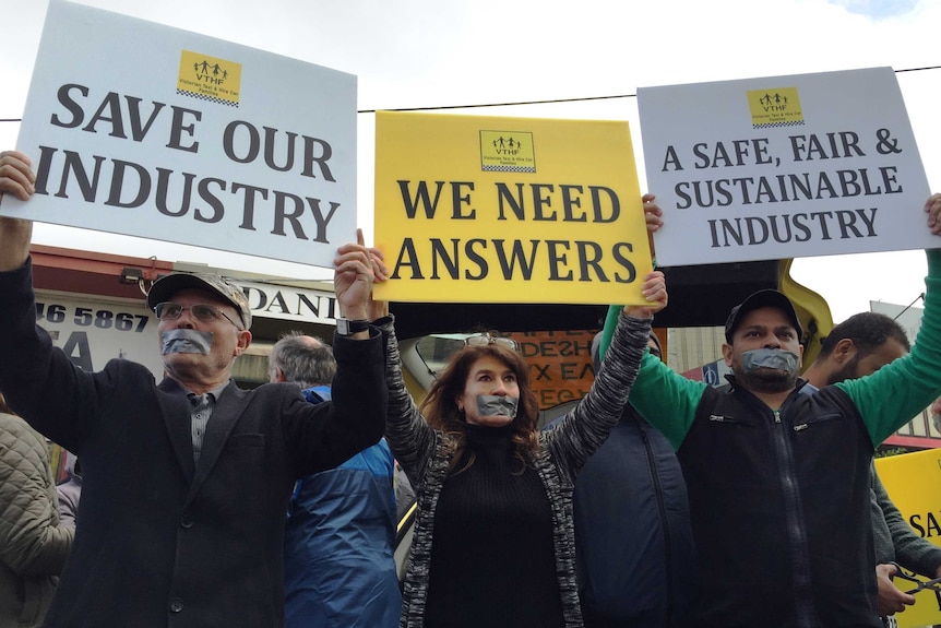 Three taxi drivers with mouths covered in tape hold up protest signs