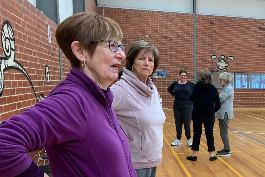 An older woman with short brown hair and glasses stands on the sideline of an indoor basketball court