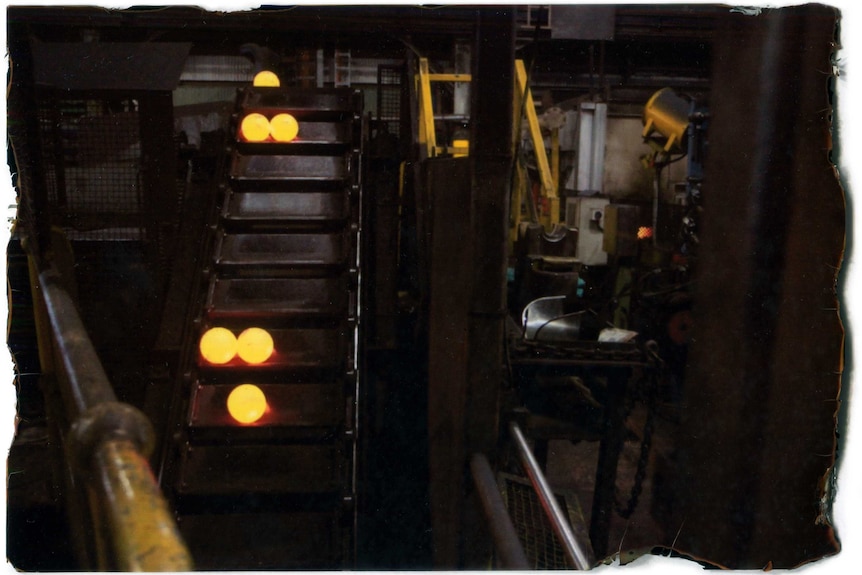 A rising conveyor belt in a factory carries hot glowing balls of steel.