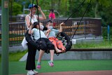 A woman pushes a swing with kids on it wearing masks.