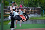 A woman pushes a swing with kids on it wearing masks.