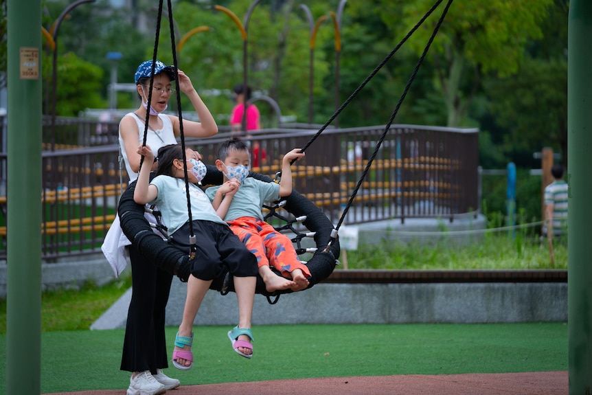 A woman pushes a swing with kids on it wearing masks.