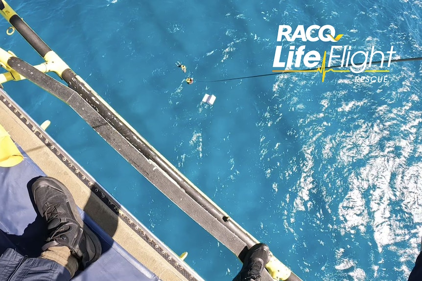 A view down at the ocean from a helicopter, of two people in the water in life jackets and an esky