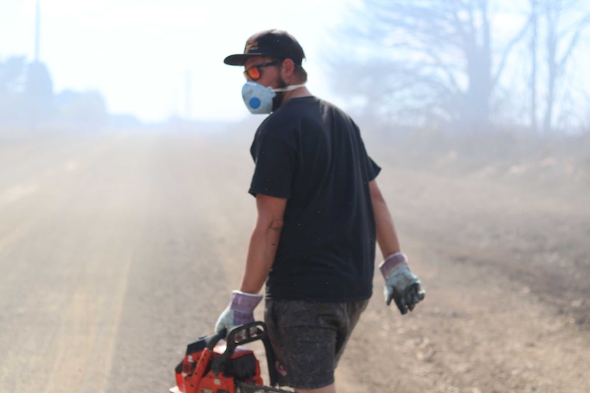 A man walks down smoky road holding a chainsaw and wearing a gas mask