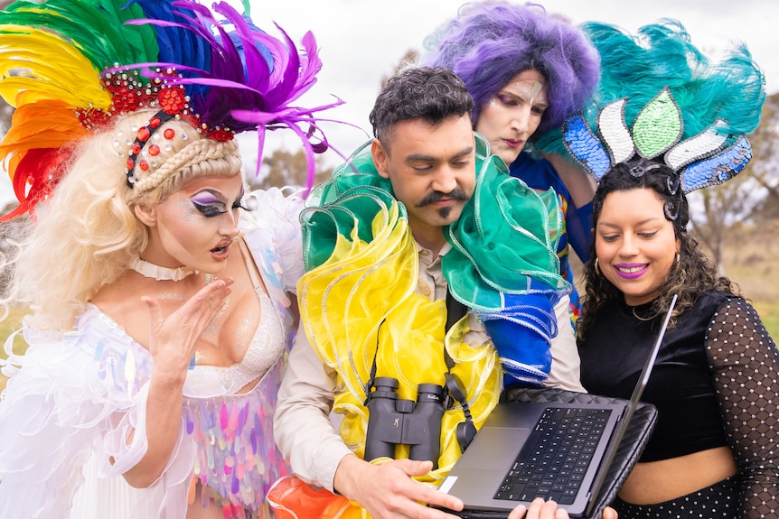 A group of elaborately dressed performers crowd around a laptop outside.